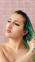 A model with winged eyeliner and facial piercings washes their wet, green-tinted hair in the shower with the Lush "Retread" conditioner. Water droplets fall around them against a background of pink square tiles.